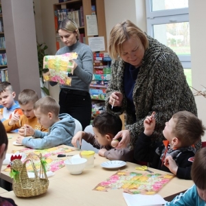 Dzieci siedzą przy stole. Dwie panioe stojące koło dzieci.