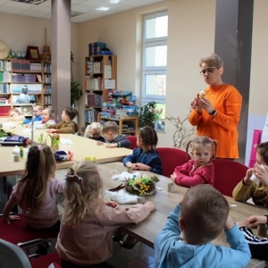 Grupa dzieci siedzą przy stole, obok stoi kobieta i objaśnia jak wykonać ozdobę wielkanocną.