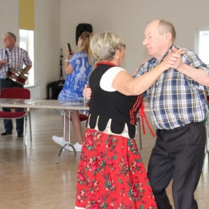 Kobieta i mężczyzna tańczy w parze , reszta uczestników spotkania przygląda się. Dwóch mężczyzn gra na instrumentach.