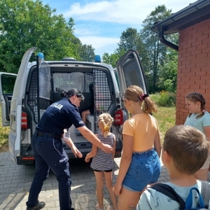 Policjantka pomaga dziewczynce wsiąść do radiowozu, troje dzieci czeka obok