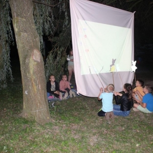 Przy dużym ekranie siedzą dzieci, z jednej strony osoby biorące udzieł w teatrzyku cieniu,a z drugiej widzowie.