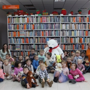 Przedszkolaki z pluszowymi misiami, z bibliotekarkami i nauczycielkami, uśmiechają się w kierunku osoby robiącej zdjęcie.