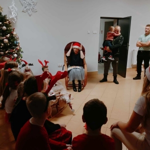 Bibliotekarka w mikołajowej czapeczce siedzi w bujanym fotelu, czyta dzieciom książkę