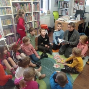 Uczestnicy spotkania siedzą w kręgu, bibliotekarka czyta im książkę