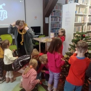 Dzieci wieszja ozdoby na choinkę, obok stoi bibliotekarka, na szyi ma zawieszony błyszczący łańcuch, dwie dziewczynki patrzą na nią z uśmiechem