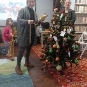 Bibliotekarka z nauczycielką stoją obok choinki, trzymają w ręku łańcuch z błyszczących koralików 