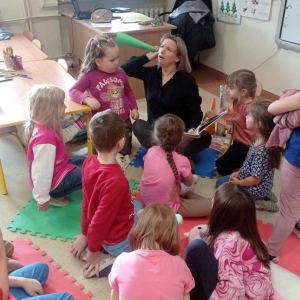 Bibliotekarka z zabawną miną trzyma papierową trąbkę przy uchu, udaje że podsłuchuje, grupa dzieci się z tego śmieje