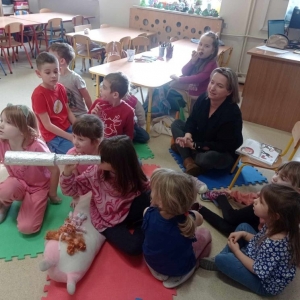 Dziewczynka spogląda przez lunetę, kilkoro dzieci i bibliotekarka zgadują na co patrzy.