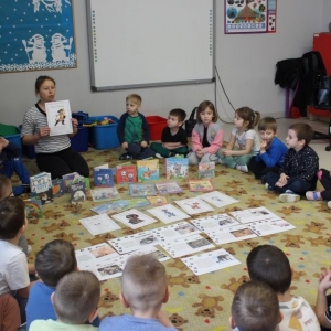 Dzieci siedzą w okręgu na dywanie przed nimi leżą rozłozone rysunki kotów i książki. Bibliotekarka prezentuje książkę.