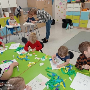 Dzieci przy stolikach oraz w krzesełkach dekorują kwiatami z papieru Panią wiosnę .