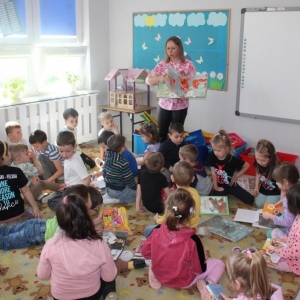 Na dywanie dzieci oglądają książki, przed nimi stoi bibliotekarka trzyma w rękach dużą kolorową książkę.