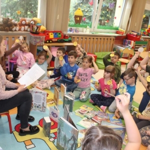 Bibliotekarka czyta zagadki, przed nią w okręgu siedzą dzieci, trzymają w rękach żółte obrazki.