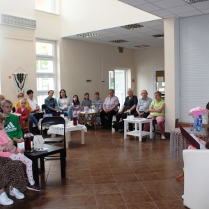 Na holu biblioteki przy stolikach siedzi grupa ludzi, przed nimi autorka czyta wiersze, wszyscy patrzą na nią z zaciekawieniem.