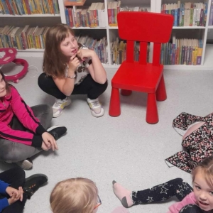 Dzieci oraz bibliotekarka siedzą na podłodze, bibliotekarka trzyma książkę.