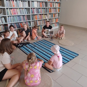 Dzieciwraz z prowadzącą siedzą na dywanie, bibliotekarka trzyma książkę.