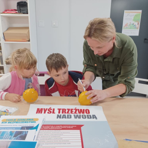 Pani bibliotekarka usuwa za pomocą kleszczołapki szpilkę z pomarańczy, dwoje dzieci się przygląda