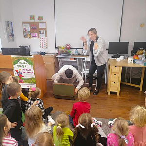 Prowadzący szuka przedmiotu w walizce, zdziwiona bibliotekarka stoi obok