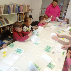 Grupa przedszkolaków pod okiem opiekunki wykonuje prace plastyczne.