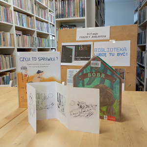 Na stoliku dwie książki i papierowa książeczka do kolorowania, w tle gazetka tematyczna - Tydzień Bibliotek.