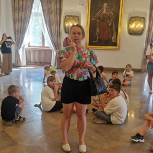 Kobietaw sali muzealnej za nią na podłodze siedzi grupa dzieci
