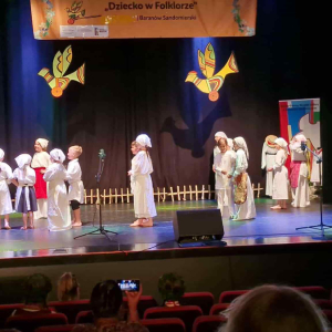 Dzieci na scenie stoją zwrócone w stronę kobiety ze skrzypcami ubranej w  strój ludowy
