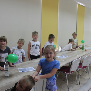 Dzieci stoją przy stole . Na stołach butelki z nałozonymi wypełnionymi balonami.