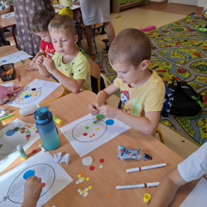 Kilkoro dzieci siedzących przy stoliku wykleja rysunek balona kolorowymi kropkami