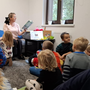 Bibliotekarka z małym, czarnym misiem na ramieniu czyta dzieciom książkę.