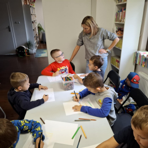 Pani opiekunka grupy rozmawia z chłopcem, kilku chłopców rysuje.