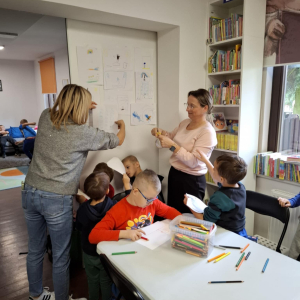 Bibliotekarką z opiekunką grupy przyklejają prace plastyczne na tablicy, kilku chłopców się przygląda.