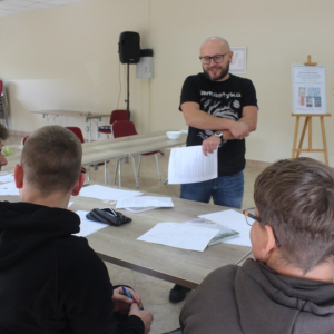 Trzej młodzieńcy siedzą przy stole przed prowadzącym