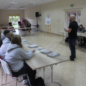 Uczestnicy siedzą przy stołach pośrodku sali stoli prowadzący