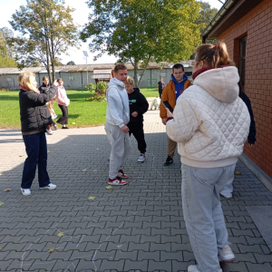 Młodzież z nauczycielką testuje prototyp gry planszowej na parkingu przed biblioteką