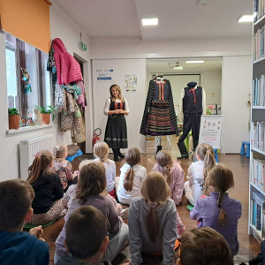 Młoda kobieta ubrana w strój ludowy opowiada o stojącej obok ekspozycji męskiego i kobiecego stroju ludowego.