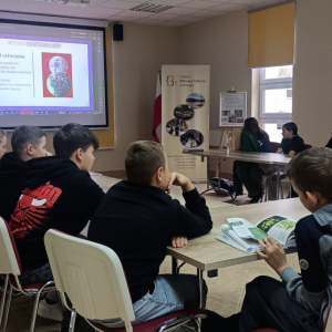 Uczestnicy siedzą przy sołe ustawionym w podkowę i oglądaja prezentację