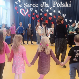 Dzieci stoją w okręgu trzymając się za ręce. W środku stoi prowadząca 