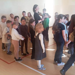 Dzieci w parach stoją po okręgu, kobieta stoi po środku koła i gra na skrzypcach, mężczyzna stoi poza okręgiem