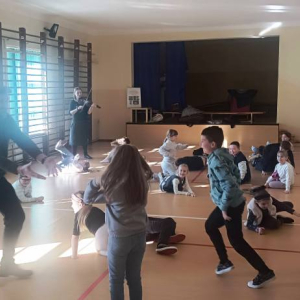 Dzieci leżą na podłodze powtarzając gesty prowadzącego warsztaty.