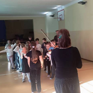 Chłopcy i dziewczynki stoją w  szpalerze prZed nimi stoi kobieta ze skrzypcami