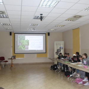 Grupa uczniów siedzi przy stołach oglądając prezentację  przed nimi stoi prowadząca warsztaty