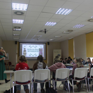 Grupa uczniów siedzi przy stołach oglądając prezentację  przed nimi stoi prowadząca warsztaty