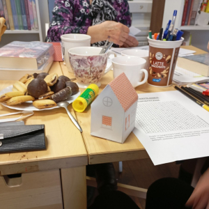 Papierowy domek, w tle stół z materiałami plastycznymi