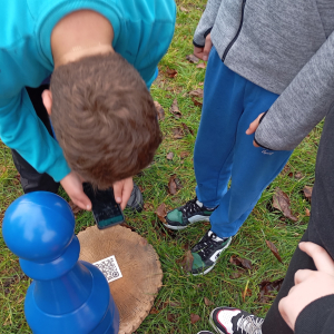 Na zdjęciu widać grupę chłopców wokół niebieskiego pionka, który znajduje się na planszy. Jeden z chłopców trzyma telefon, skanując kod QR, co sugeruje, że gra łączy tradycyjne elementy z nowoczesną technologią.