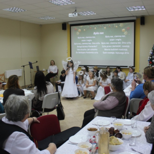 Uczestnicy wydarzenia siedząc przy stołach słuchają dziewczynki śpiewającej kolędę