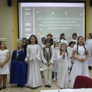 Grupa dzieci w strojach aniołków, pastuszków i króla stoją przed dużym ekranem