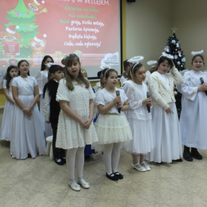 Grupa dzieci w strojach aniołków, pastuszków i króla stoją przed dużym ekranem