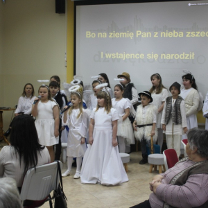 Grupa dzieci w strojach aniołków, pastuszków i króla stoją przed dużym ekranem