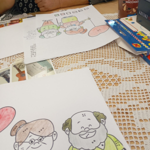 Dwa częściowo pokolorowane obrazki z babcią i dziadkiem, obok kredki i kostka do gry