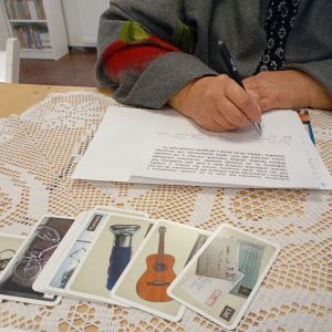 Dłonie seniorki zapisujące tekst na kartce, przed nią rozłożone karty z obrazkami