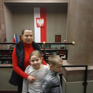 Zdjęcie uczestniczki wycieczki z dwójką dzieci w przestrzeni Sejmu, na tle godła państwowego.